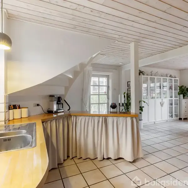 Et køkken med en buet træbordplade indeholder vask og en kaffemaskine. Køkkenet åbner op til en lys stue med hvidt flisegulv, spisebord og vitrineskab.