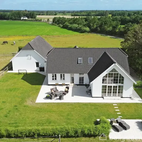 Hvidt, moderne hus med mørkt tag og store vinduer står omgivet af en grøn have. Solterrasse med møbler foran huset. Mark og skov i baggrunden. Ingen synlig tekst.