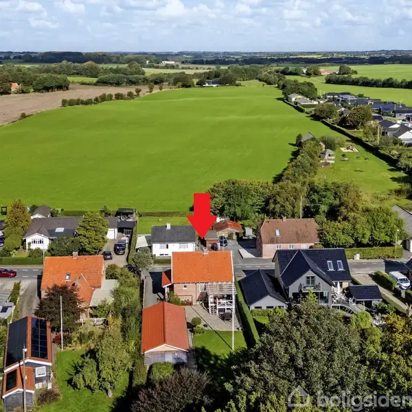 Et hus, markeret med en rød pil, står blandt andre huse i et landligt område omgivet af grønne marker og en spredt beboelse i baggrunden.