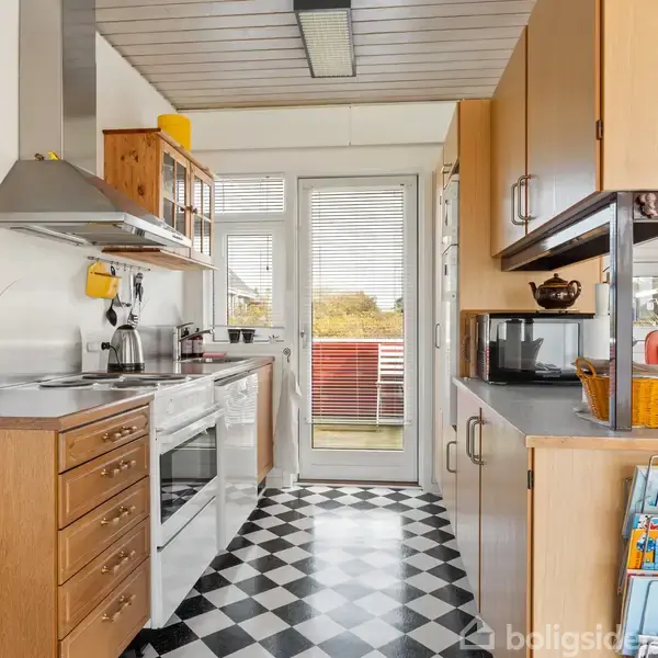 Køkken med træskabe og køkkengrej; rummet har et ternet sort-hvidt gulv og en glasdør fører til en balkon. Et reolsystem indeholder tidsskrifter og en mikrobølgeovn.