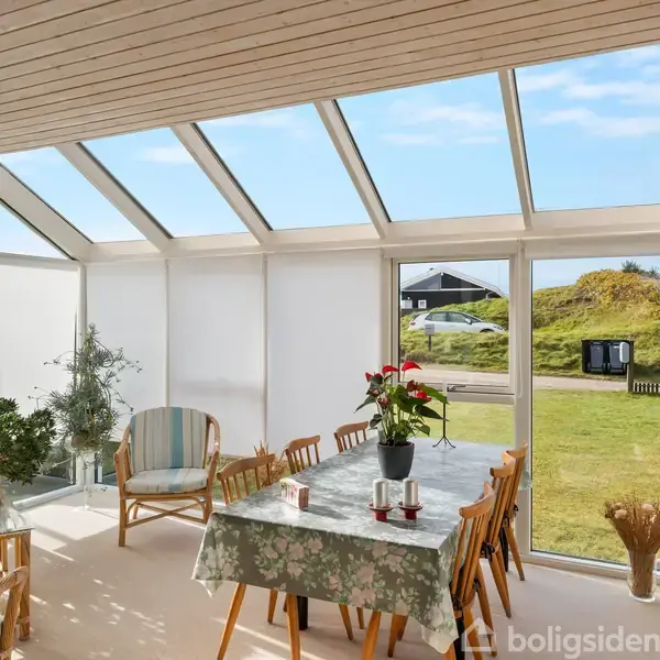 Spisestue med glasvægge viser stort bord dækket med blomstrede dug og blomster, omgivet af stole. Udenfor ses en solrig dag med grønne bakker og huse i det fjerne.