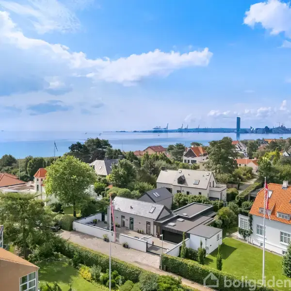 Huse med røde tage står stille i en frodig, grøn villakvarter nær kysten, med havet og en industrihavn synlig i horisonten under en let overskyet, blå himmel.
