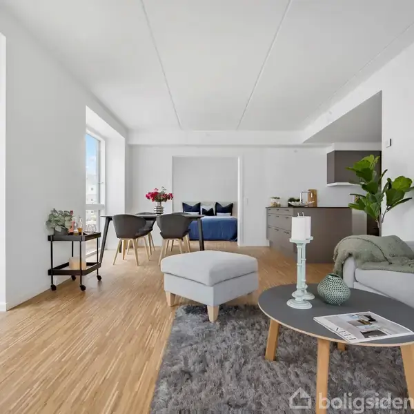 Stue med sofa, lænestol, sofabord og tæppe. Ved spisebordet står fire stole. Rummet har lyse vægge, store vinduer og trægulve. Plante i hjørnet. Lys og moderne indretning.