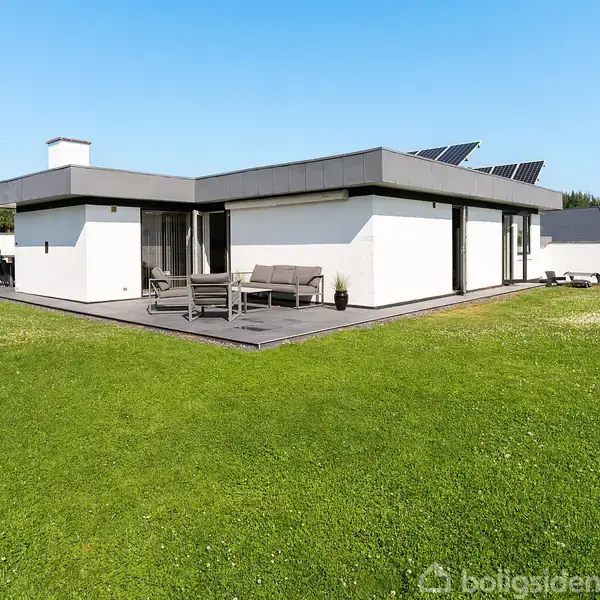 En moderne, hvid bungalow med fladt tag og solpaneler står i en velplejet grøn have. Der er en terrasse med havemøbler. Himlen er klar og blå.