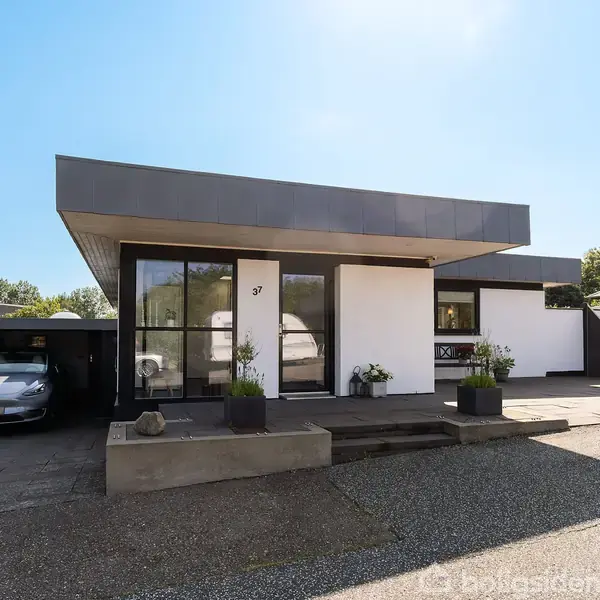 En moderne, hvid murstensvilla med fladt tag, står stille i et solrigt boligområde. Foran er nummeret 37 synligt. En indkørsel rummer en bil i carporten til venstre.