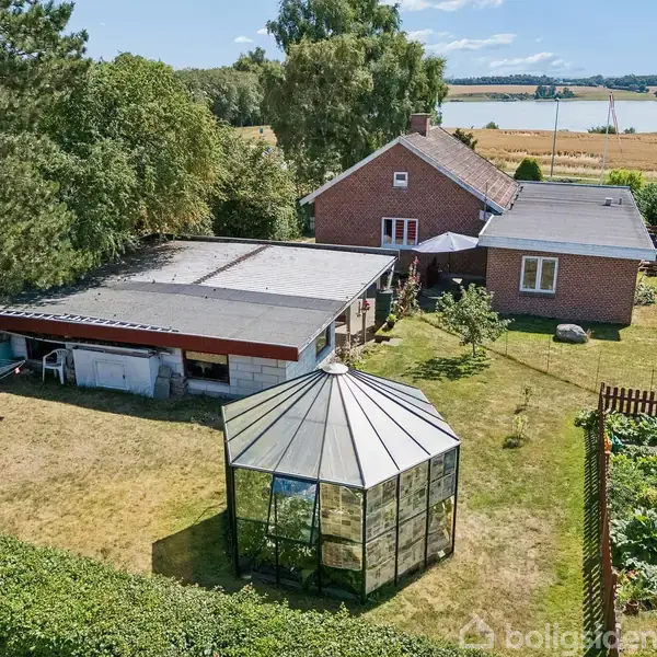 En ottekantet pavillon står i en grøn have med et drivhus, omgivet af en græsplæne. I baggrunden ses et rødt murstenshus med udsigt over marker og vand.