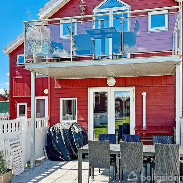 Rødt hus med altan øverst, glasrækværk og havemøbler på terrassen. Solskin og blå himmel skaber en lys atmosfære omkring hjemmets udendørsområde. Poletter omkring terrassen tilføjer privatliv.