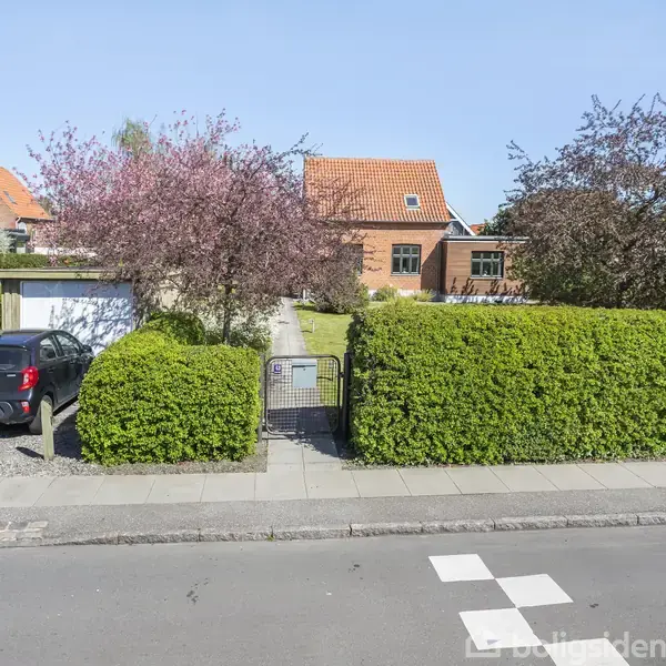 En bil holder parkeret ved siden af en garage omgivet af grønne buske. En orange murstensbygning med rødt tag ses i baggrunden, omgivet af blomstrende træer og en flisebelagt sti.