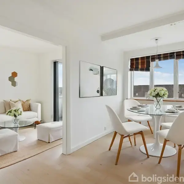 Spisestue med rundt bord, fire hvide stole og blomsterdekoration, beliggende ved siden af stue med sofa og sofabord. Store vinduer giver udsigt og naturligt lys i begge rum.