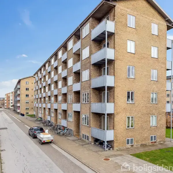En række lejlighedsbygninger i gule mursten strækker sig langs en vej. Cykler parkeret foran bygningen. Biler parkeret til venstre på vejen. Blå himmel ovenover.