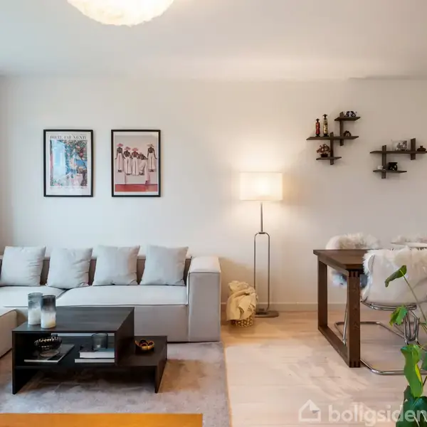 Sofaen står ved en væg med to indrammede billeder, ved siden af et sofabord. I hjørnet er der en gulvlampe. Stuen rummer også et spisebord og en plante.