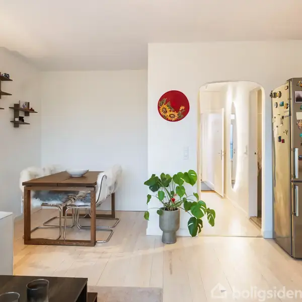 Et moderne køkken-alrum med et træbord, omkranset af to stole i en hyggelig spisekrog. Til højre står et køleskab dekoreret med magneter. En plante står ved siden af.