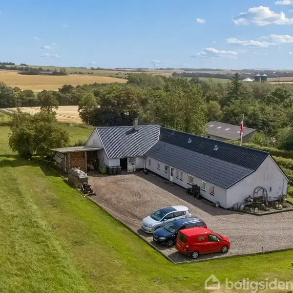 En lang, hvid bygning med sort tag står i en grusindkørsel. Tre biler parkeret foran. Omkringliggende vidder med marker og træer omfavner landskabet under en blå himmel med skyer.