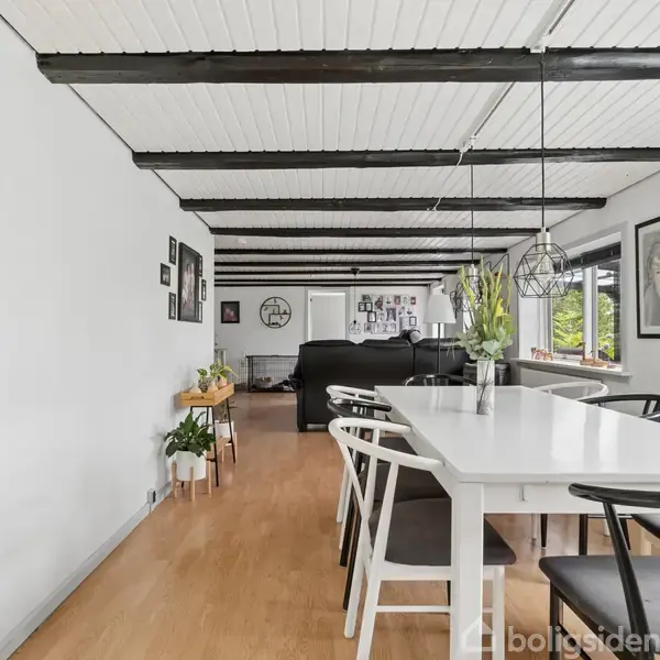 Et hvidt, rektangulært spisebord står midt i et lyst rum med sorte stole omkring. På bordet er en vase med planter. I baggrunden ses en stue med sofaer og vinduer.