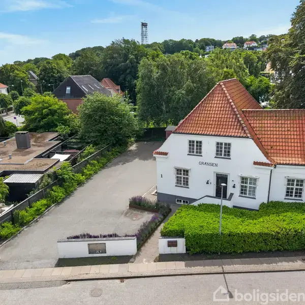 Et hvidt hus med rødt tegltag står på en rolig gade. Skiltet "GRÆNSEN" ses foran huset. Omgivet af grønne træer og en mindre indkørsel.