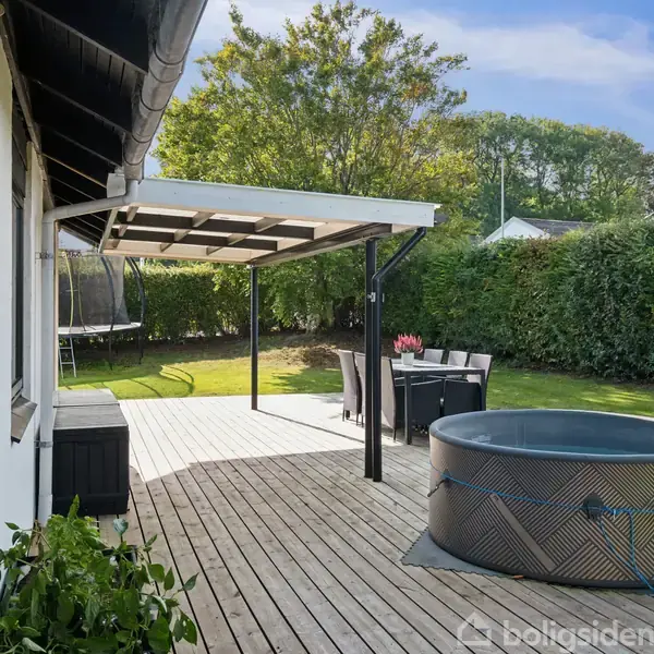 Husets terrasse med trædæk har et bord med seks stole under et halvtag og et rundt udendørs bassin. Græsplæne og hække omgiver området, og træer ses i baggrunden.