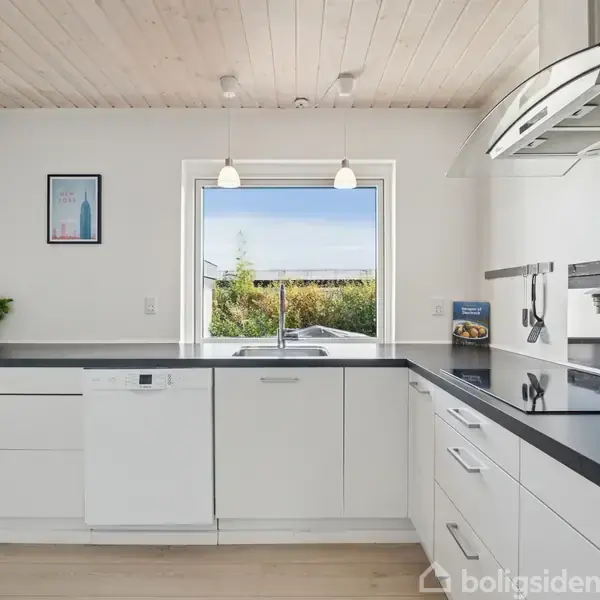 Et lyst køkken med hvide skabe og sort bordplade. En opvaskemaskine er indbygget, og der er en stor vask under et vindue. En plante og en plakat ses til venstre.