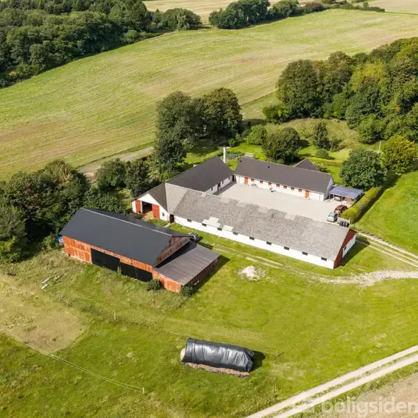 En gård med tre bygninger ligger i en landlig, grøn eng omgivet af marker og skov. Der er en grusvej, der fører til gården, omkranset af træer.