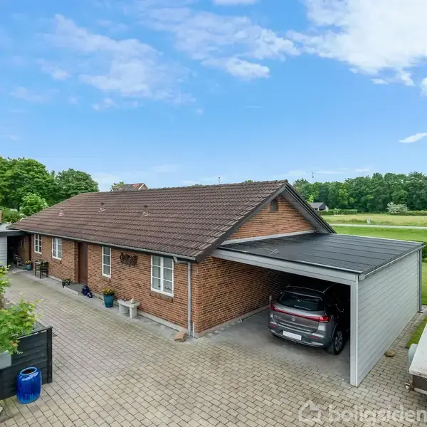 En etplans villa med brun murstensfacade står stille på en flisebelagt indkørsel. I carporten er en bil parkeret. Græsplæne og træer omgiver huset, og flag er i baggrunden.