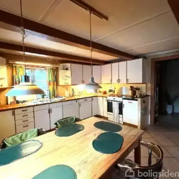 Et køkken med et langbord dækket af fire grønne dækkeservietter, omgivet af stole. Køkkenet har hvide skabe, en ovn, køleskab og hængende lamper. En dør leder til et andet rum.