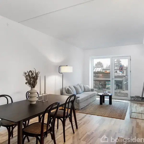 En moderne stue med en grå sofa foran et stort vindue, et sort spisebord med fire stole og en gulvlampe. Rummet har lyst trægulv og hvidmalede vægge.