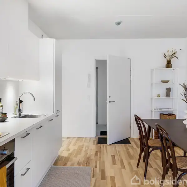 Et moderne køkken-alrum med hvide skabe fyldt med køkkenredskaber, en åben dør i baggrunden, og et spisebord med stole, omgivet af lyse trælaminatgulve.