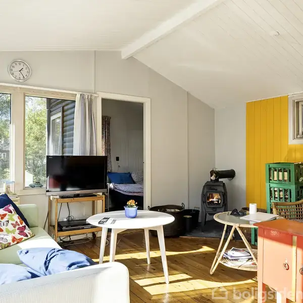 En lys dagligstue med en sofa fyldt med farverige puder, hvor et TV står på et lille bord foran vinduerne. En brændeovn står ved en gul væg.