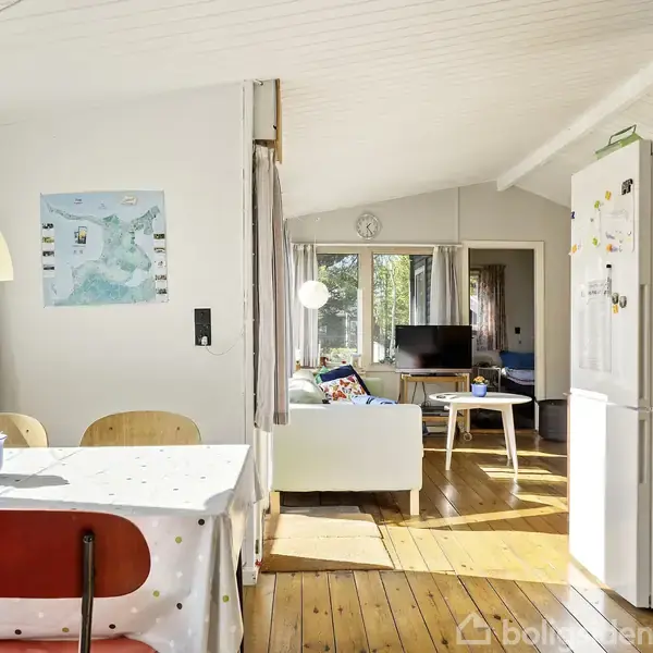 Et lyst køkken-alrum med spisebord dækket af en dug, dekoreret med en vase med blomster. Til højre står et køleskab og træmøbler. I baggrunden ses en stue med sofaer.