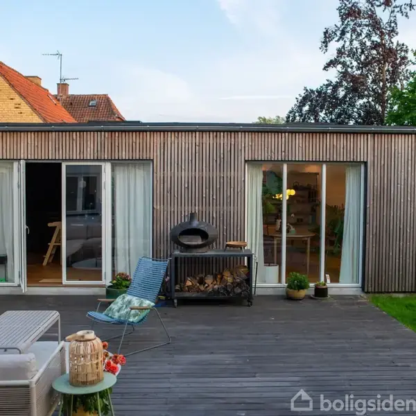 En moderne, enkel bygning med træbeklædning har skydedøre, der åbner mod en terrasse med havemøbler. I baggrunden ses grønne træer og nabohuset med en gul murstensfacade.