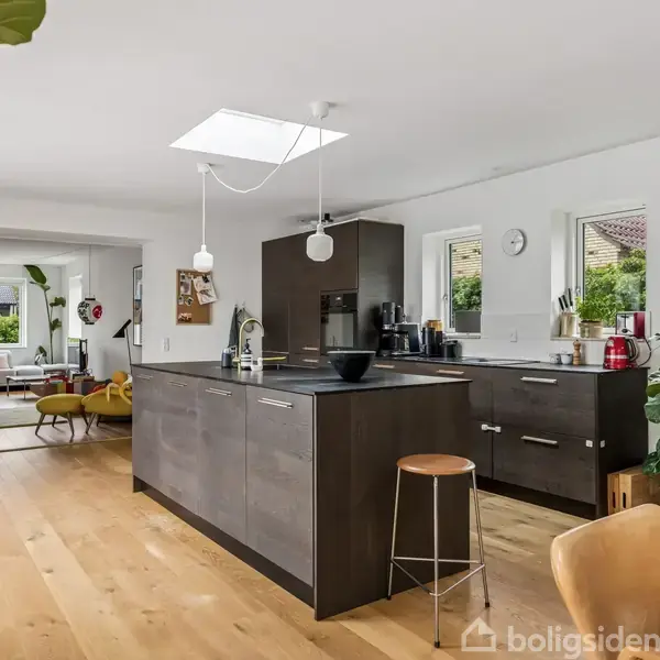 Et moderne køkken med mørke skabe og en ø-bar, hvorpå der står køkkenredskaber. I baggrunden ses en stue med sofaer og store vinduer, der lukker lys ind.