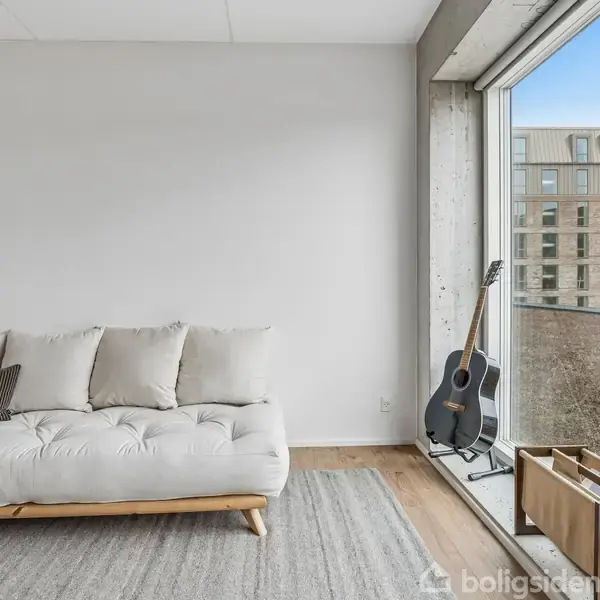 En lys stue med en hvid sofa placeret på et tæppe. Ved siden af står en akustisk guitar op ad vinduet, som viser en udsigt over en murstensbygning.