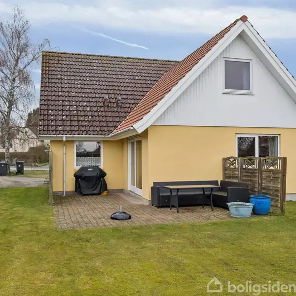 Hus med gul facade og rødt tag står stille med en terrasse foran; omgivet af grøn græsplæne og hegn. Havemøbler og krukker er placeret på terrassen.