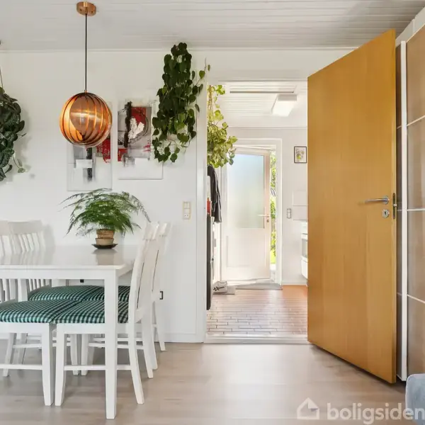 Spisebord med hvide stole står i lys stue med grønne planter og en orange lampe. En åbentstående trædør fører til en entré med dagslys.