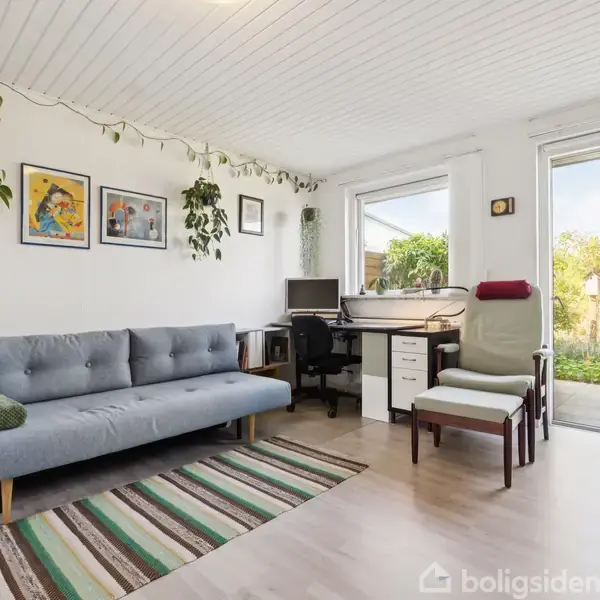 Grå sofa står på et stribet tæppe, omgivet af planter og vægkunst. Ved siden af er et skrivebord med computer. Store vinduer åbner til en have udenfor.