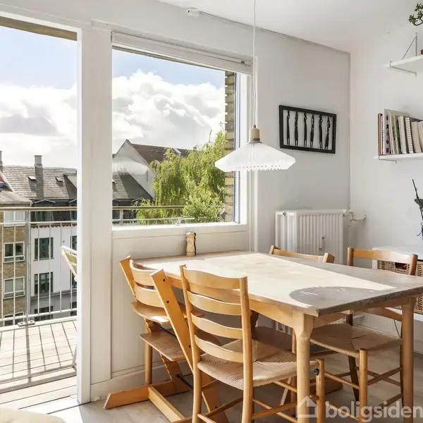 Et lyst køkken med et træbord omgivet af seks stole. Bordet er placeret ved et åbent vindue, der fører ud til en balkon med udsigt over bygninger. En hylde med bøger og planter hænger på væggen.