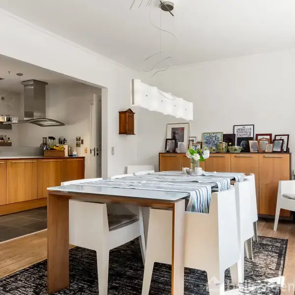 Et spisebord med hvidtækkede stole er centralt placeret i en spisestue. Bordet har en blå-hvid stribet dug. En åben køkkenadgang ses til venstre med træskabe.