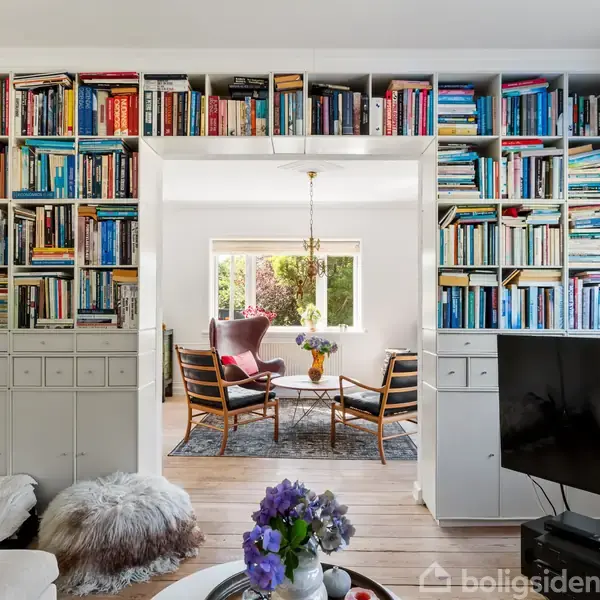 Bøger fylder reolskabe omkring en døråbning, der leder til en stue. Stuen har en lænestol, to stole, et tæppe, sofabord og en loftslampe. Forgrund: fjernsyn og blomster.