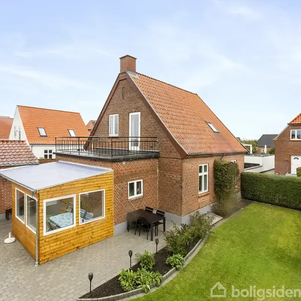 Rødt murstenshus med rødtag, terrasse og balkon omringet af et velplejet grønt område. Ved siden af er en udestue og en terrasse med havemøbler.