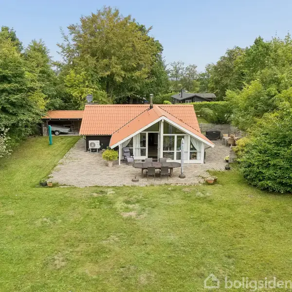 Huset med rødt tag og store vinduer står omgivet af en grøn have med tæt bevoksning. En terrasse med havemøbler ligger foran huset, omgivet af græsplæne og træer.