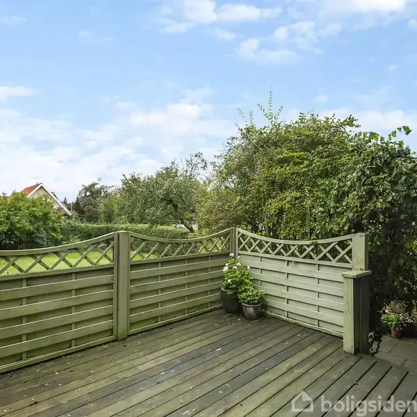 En træterrasse omgivet af et hegn og planter står i en have med græs og træer under en blå himmel.