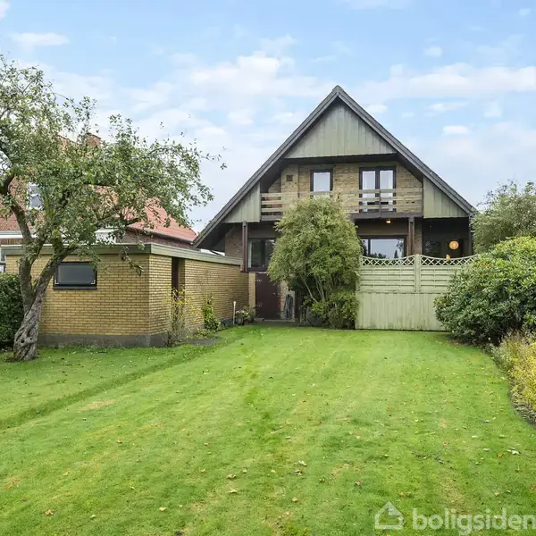 En enkeltstående, to-etagers træhus med balkon står foran en velplejet græsplæne, omgivet af hække og buske. Himmel er let skyet.