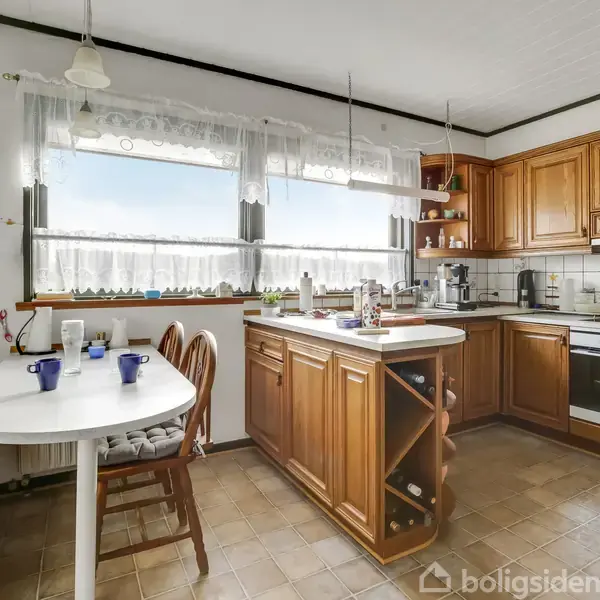 Et køkken med træskabe og en central ø, bordflader fyldt med køkkenudstyr. En hvid spisebord med blå og hvide kopper ved siden af en bænk. Store vinduer slipper lys ind.