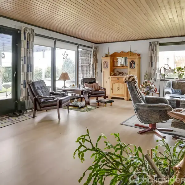 Stue med træloft; to lænestole og et bord står ved glasdøre med gardiner, som fører ud til en have. En reolvitrine står ved væggen. Rummets dekorationer inkluderer planter og lamper.