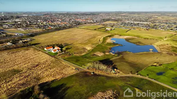Luftfoto viser en stor grøn mark med en snoet vej ved siden af en sø. Et hvidt hus med orange tag ses til venstre. En by ligger i baggrunden.