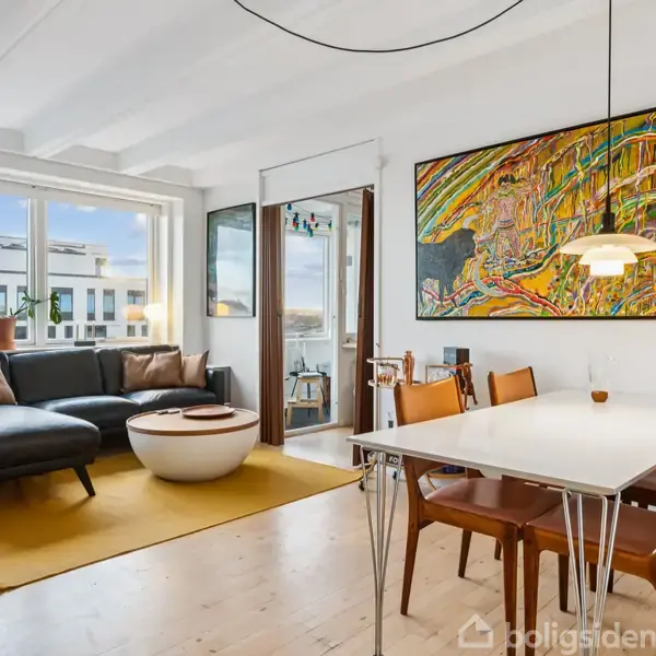 Stue med sort sofa og rundt bord på gulvtæppe, sollys strømmer gennem vinduerne. Lys spisebord med stole til højre. Farverigt maleri hænger på væggen over spiseområdet.