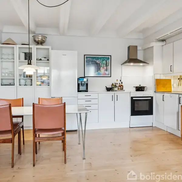 Et moderne køkken med hvidt interiør og indbyggede apparater, som omfatter en ovn og emhætte. Ved siden af står et spisebord med brune stole. Kunst og bøger pynter rummet.