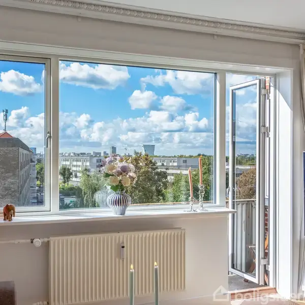 Et stort vindue åbner til en balkon med udsigt over en by under en blå himmel med skyer. Indenfor er der en vase med blomster og flere stearinlys.