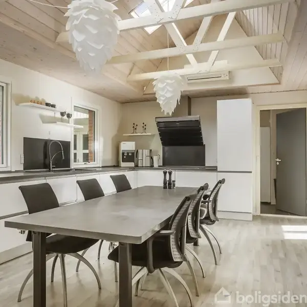 Et moderne køkken har en lang spisebord med seks sorte stole. Lyse trælofter med ovenlysvinduer skaber en lys atmosfære. Hylder og køkkenskabe omkranser rummet.