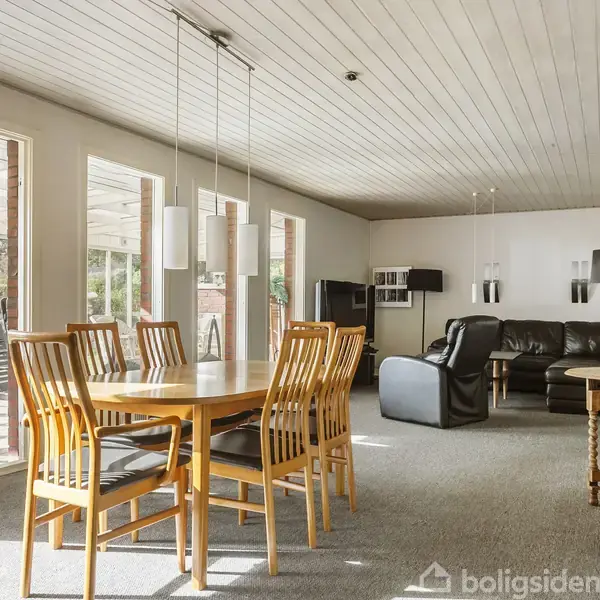 Spisebord med seks stole står i en lys stue. Sofa og lænestole i baggrunden ved glasdøre, som åbner til terrasse. Lys træloft og vægge skaber en rumlig atmosfære.