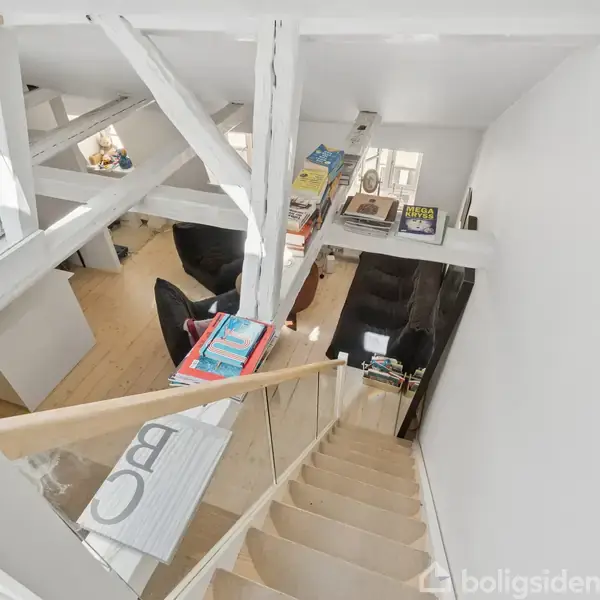Trappe fører op til en lys hems med åbent rum, hvor der er stakke af bøger på en reol. Træbjælker og store skråvinduer giver lysindfald.